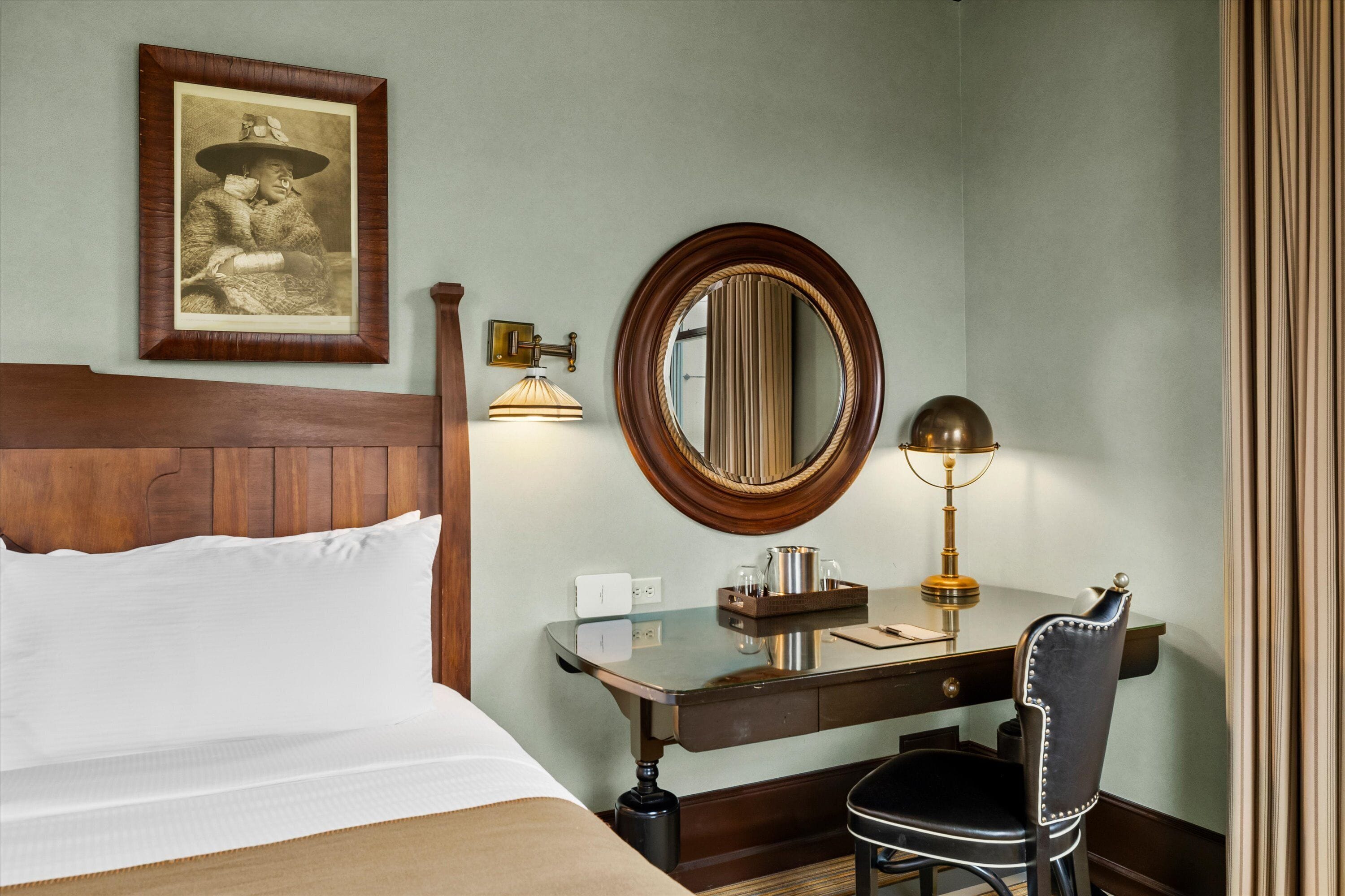 Ein Hotelzimmer mit hölzernem Kopfteil, einem Bett mit weißer Bettwäsche, einem Schreibtisch mit Lampe, einem Sessel und einem gerahmten Bild an der Wand.