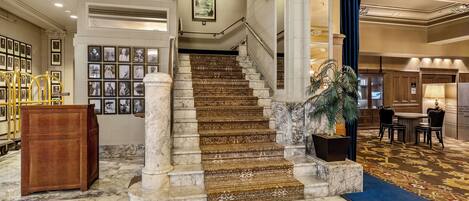 Una imponente escalera de mármol con columnas, que conduce a un rellano del segundo piso con fotografías enmarcadas.