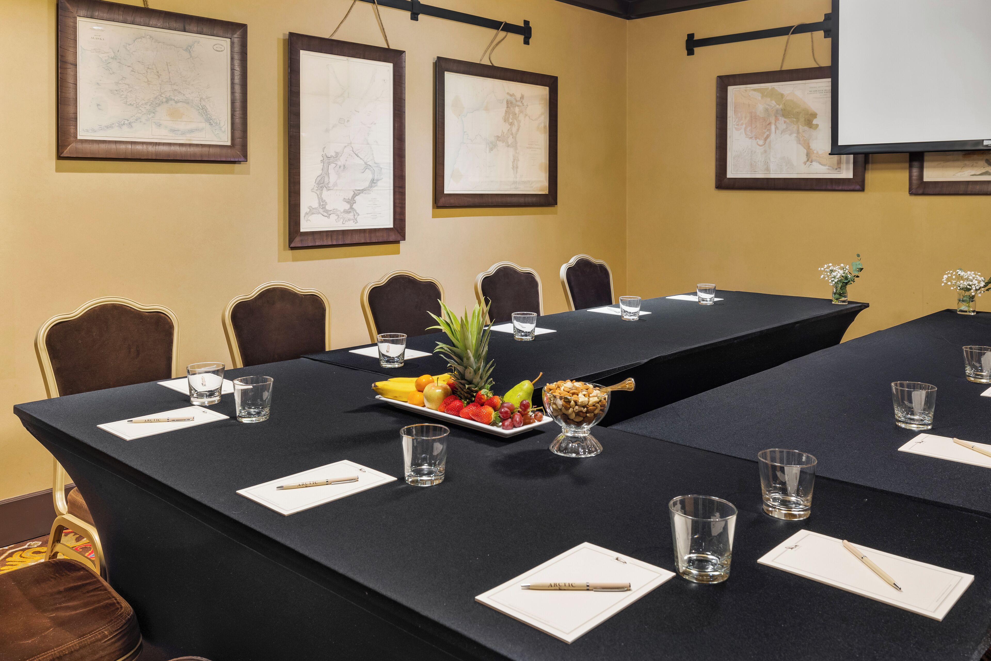 A conference room with a long table, chairs, framed maps on the wall, and a projector screen.