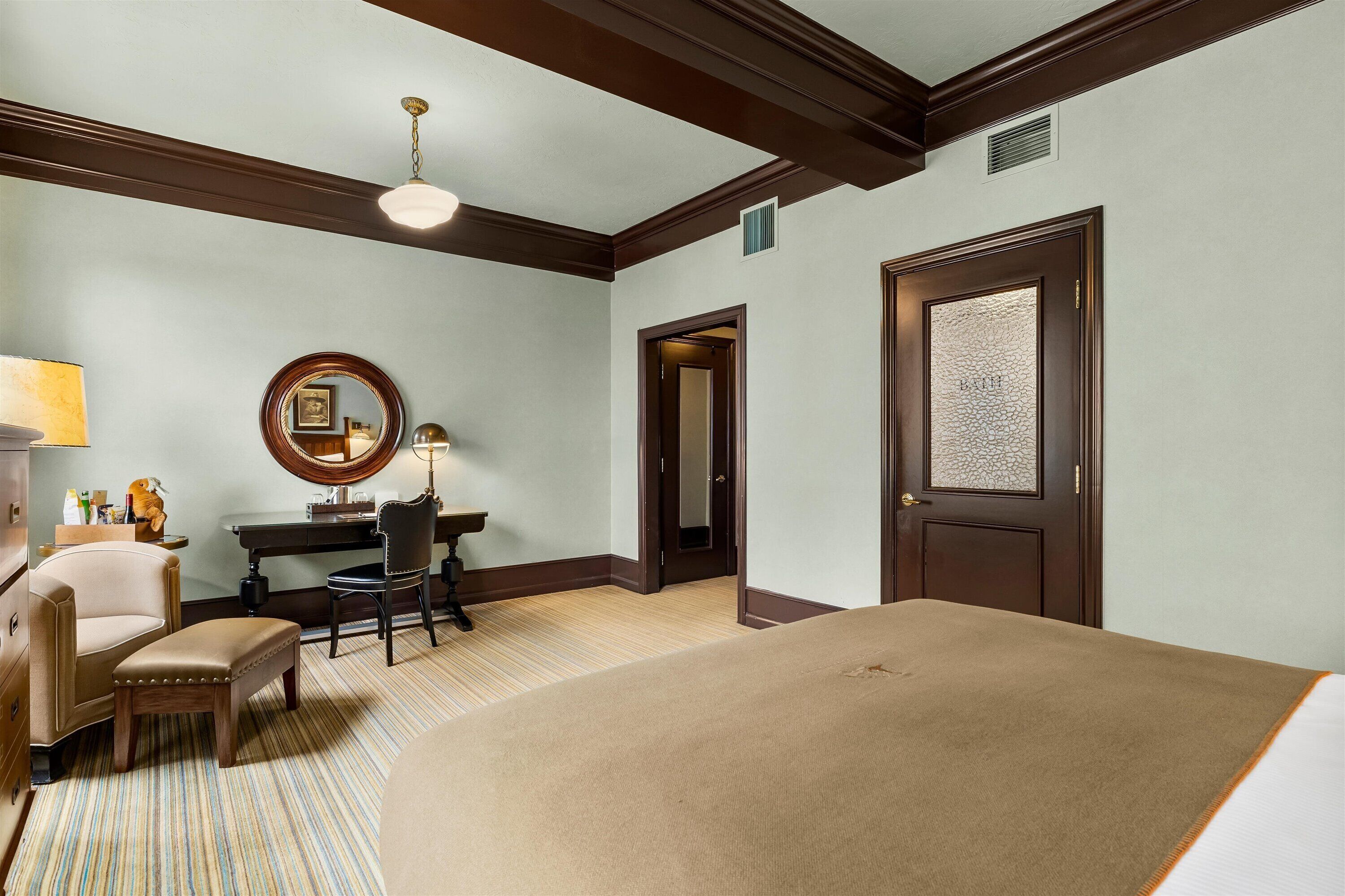 A hotel room with a large bed, a desk with a chair, a round mirror, and a lamp.