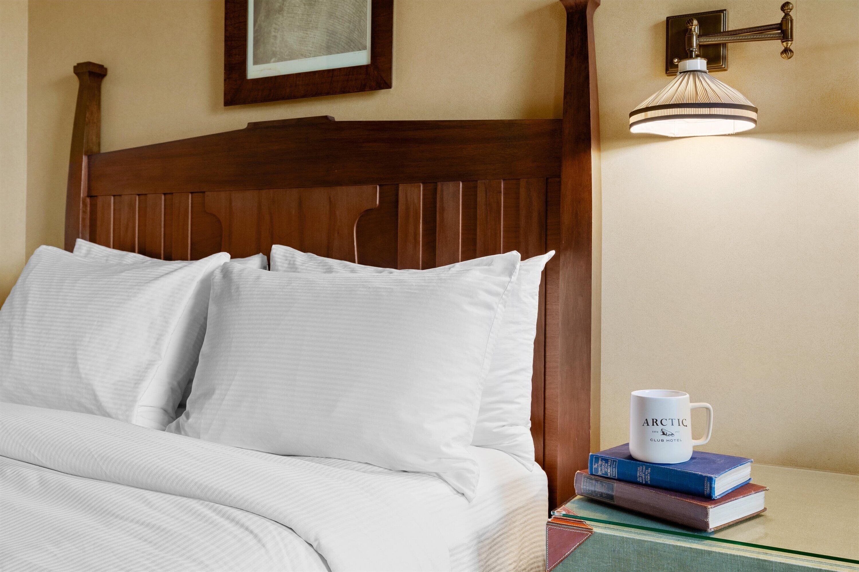 A neatly made bed with white linens, a wooden headboard, and a bedside table with a white mug and books.