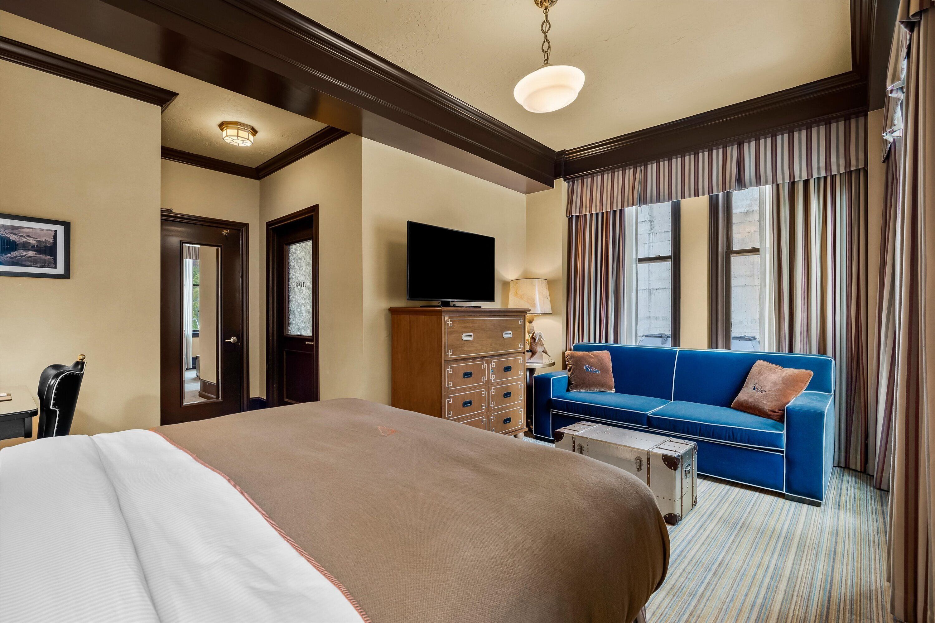 A hotel room with a large bed, a blue sofa, a television, a dresser, and a window with curtains.