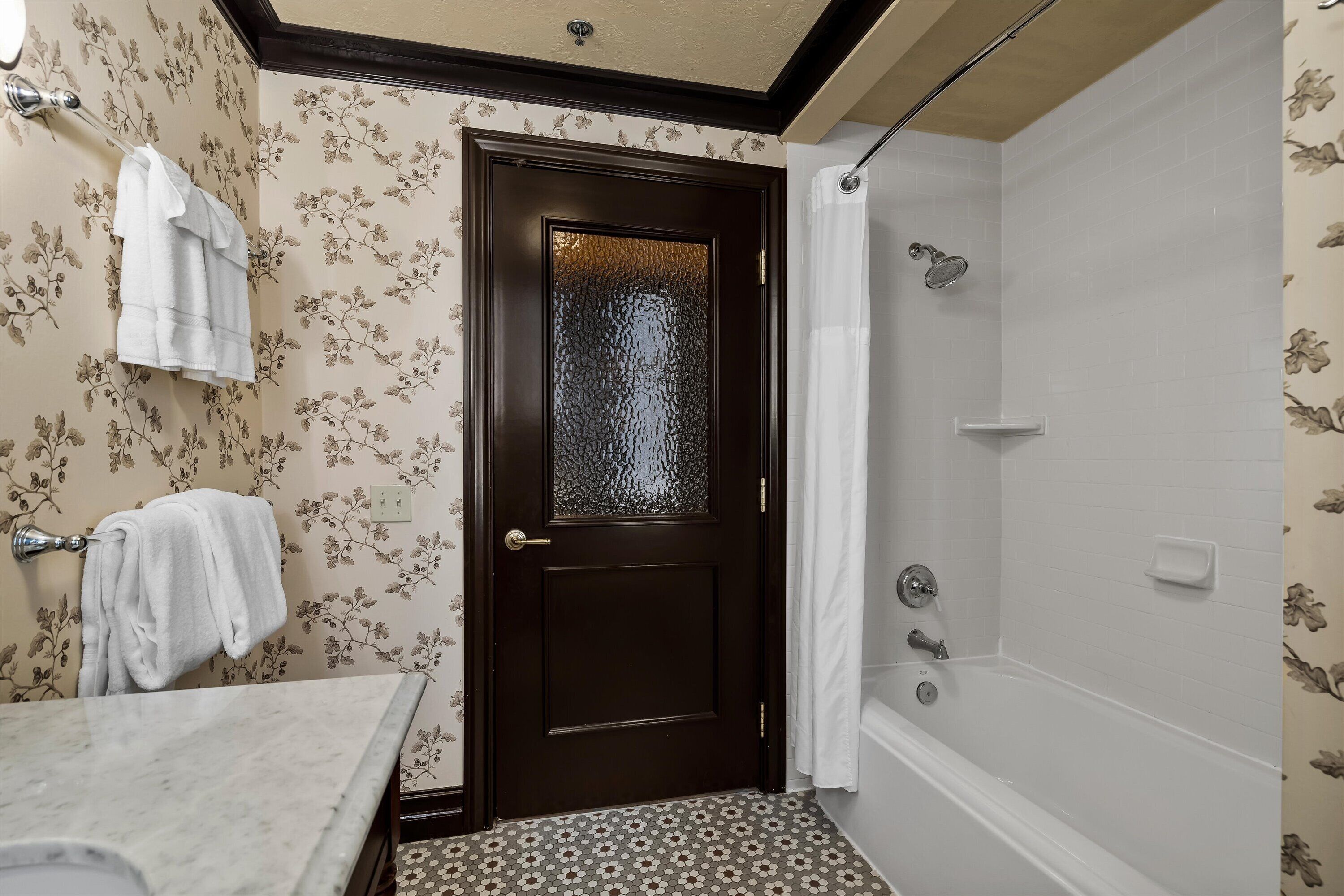 A bathroom with a dark wooden door, a shower with a glass enclosure, and patterned wallpaper.