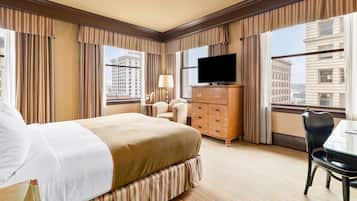 A hotel room with a large bed, a flat-screen TV, a dresser, a chair, and a view of city buildings.