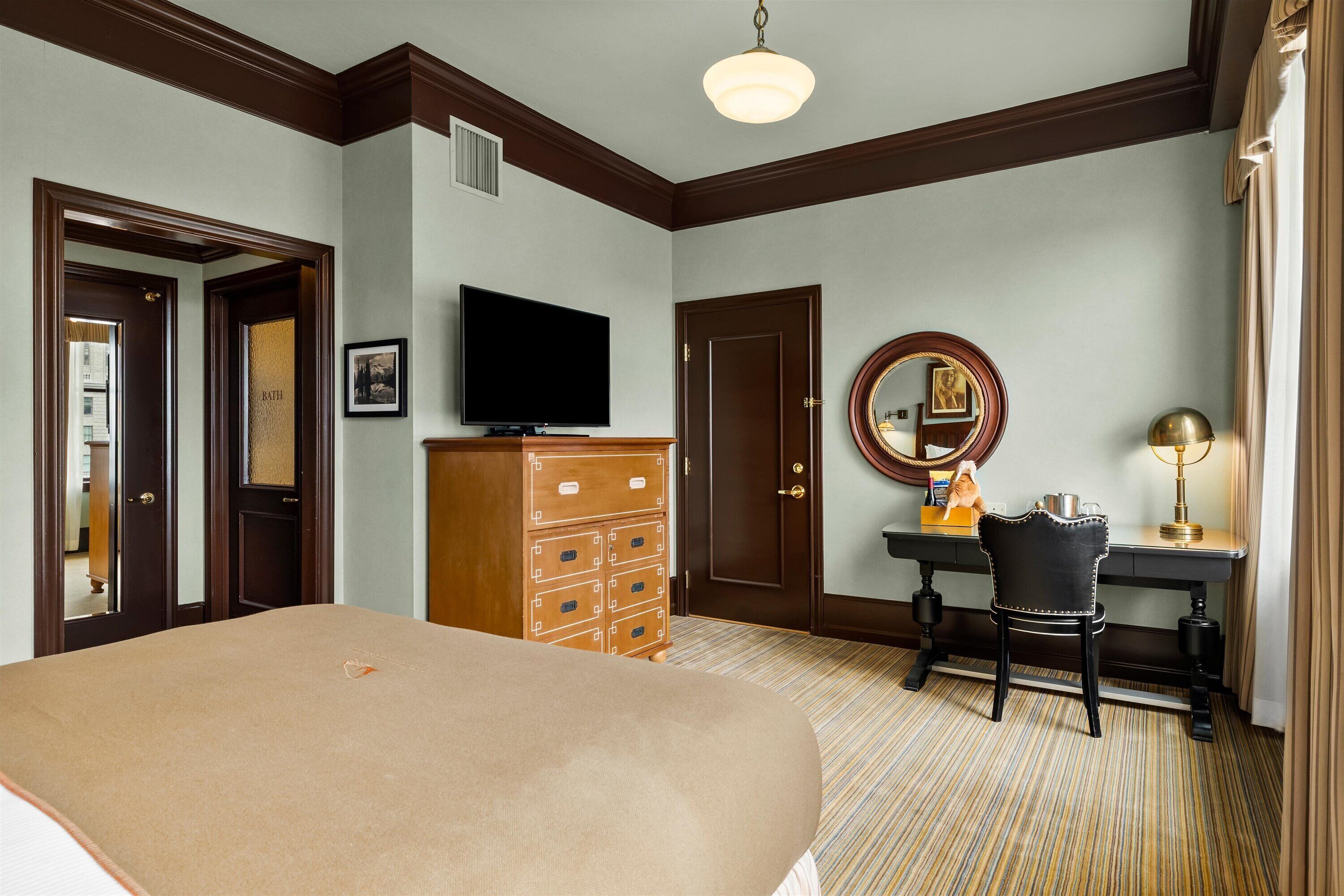 A hotel room with a large bed, a wooden dresser, a desk with a chair, a television, a mirror, and a lamp.