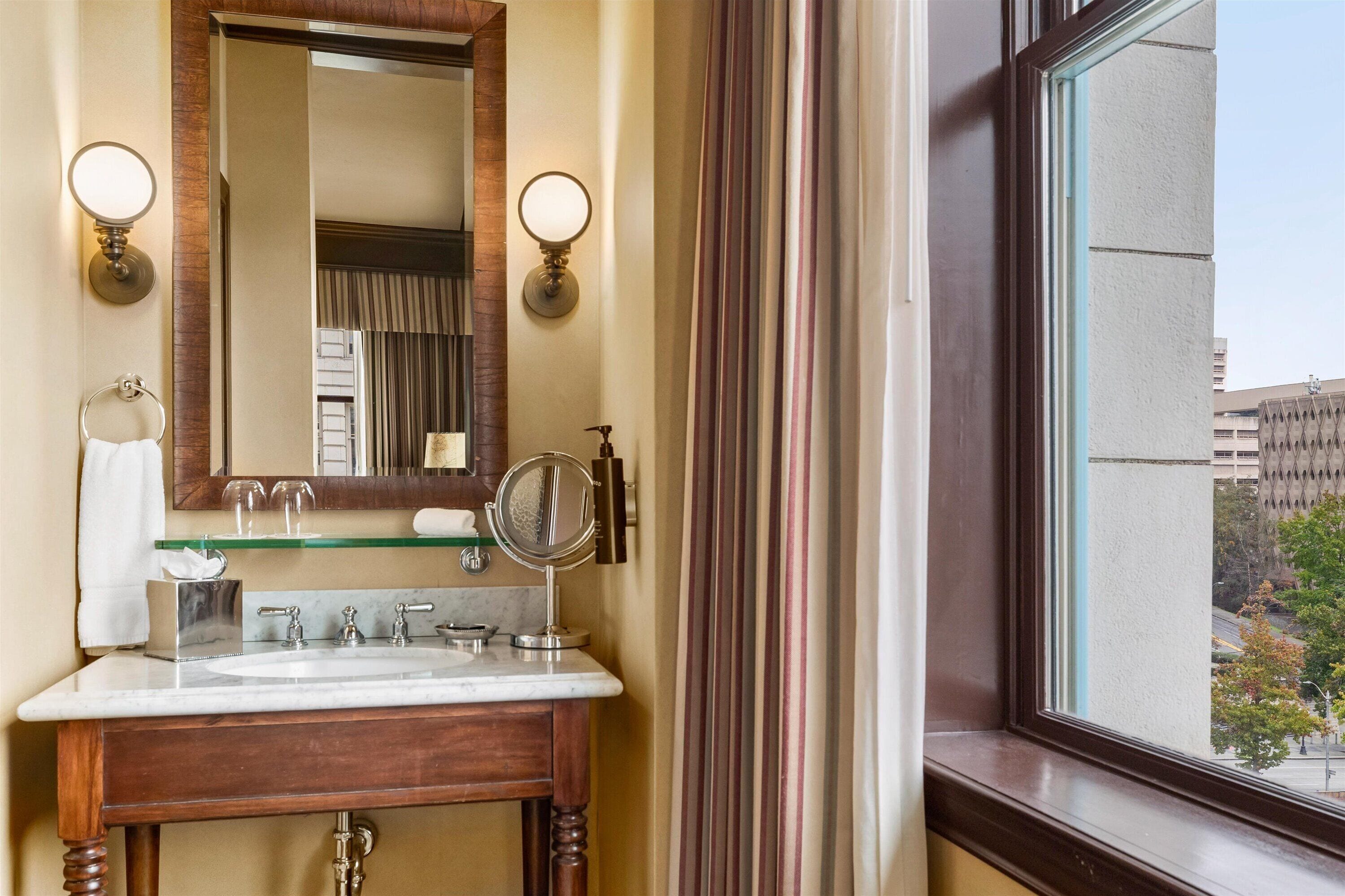 Une salle de bain avec un meuble-lavabo en bois, un miroir, deux appliques murales, un porte-serviettes et une fenêtre donnant sur des bâtiments et des arbres.