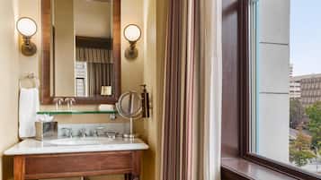 Un bagno con un mobile lavabo in legno, uno specchio, due luci a parete, un porta asciugamani e una finestra con vista su edifici e alberi.