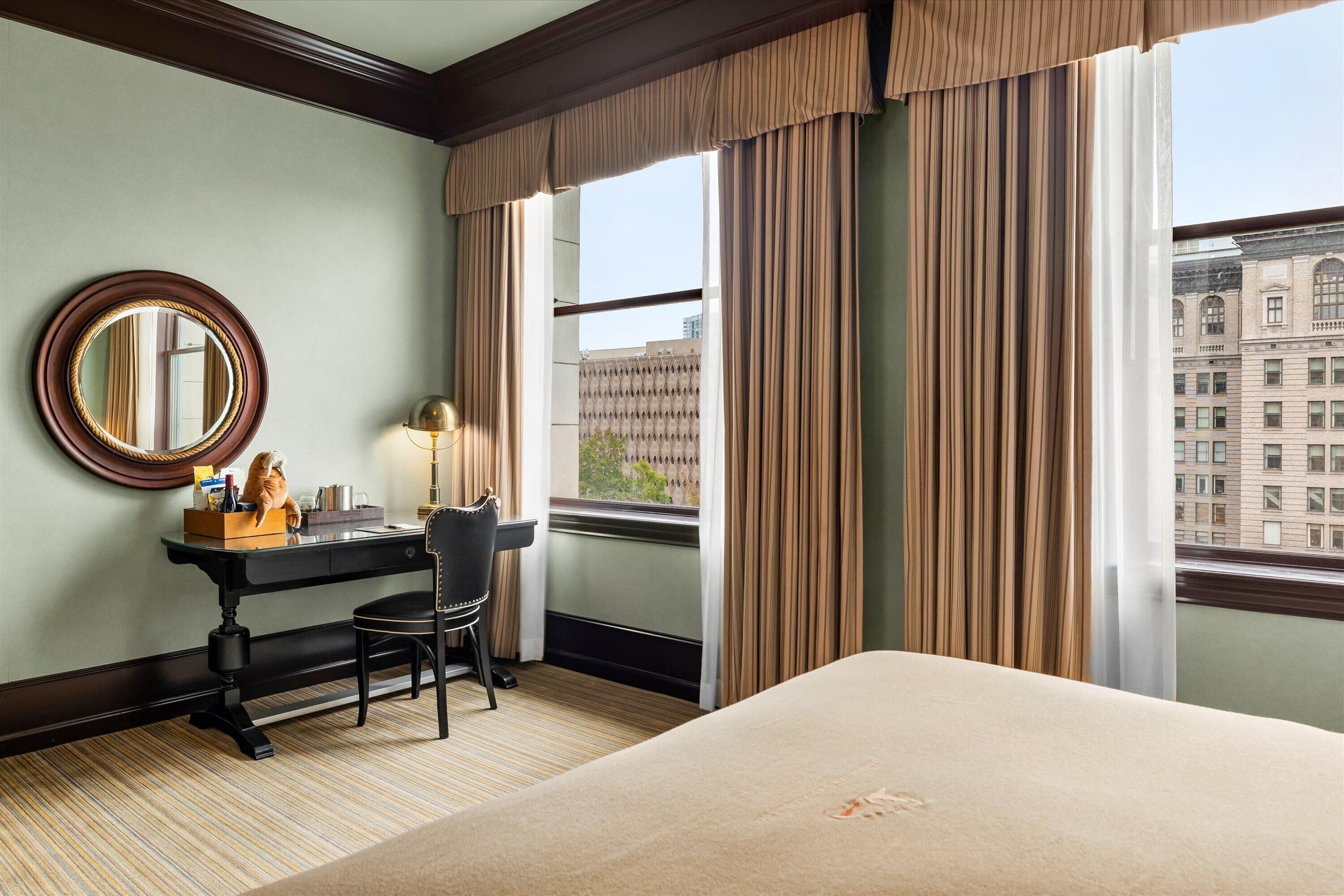 Ein Hotelzimmer mit einem großen Bett, einem Schreibtisch mit Stuhl, einem runden Spiegel und einem Fenster mit Vorhängen.