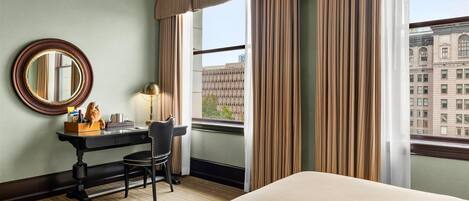 A hotel room with a large bed, a desk with a chair, a round mirror, and a window with curtains.