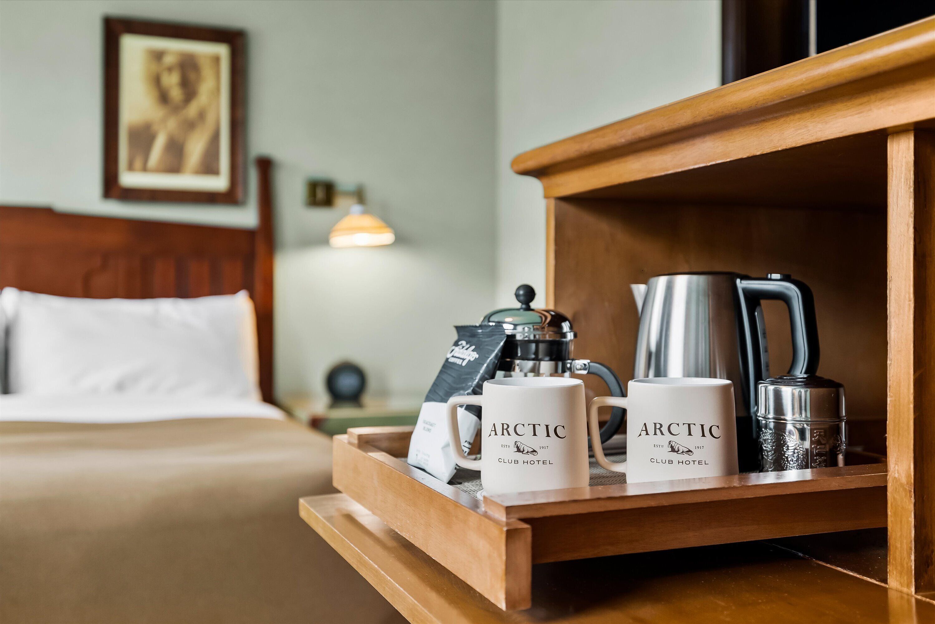 A wooden tray with two white mugs labeled "Arctic Club Hotel," a silver coffee pot, and a silver electric kettle on a hotel bed.