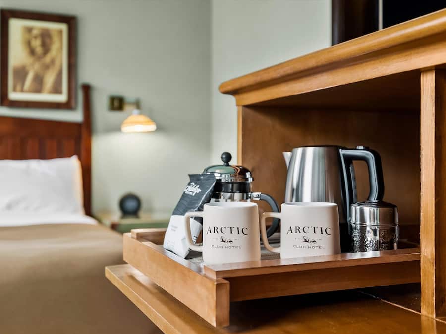 A wooden tray with two white mugs labeled "Arctic Club Hotel," a silver coffee pot, and a silver electric kettle on a hotel bed.