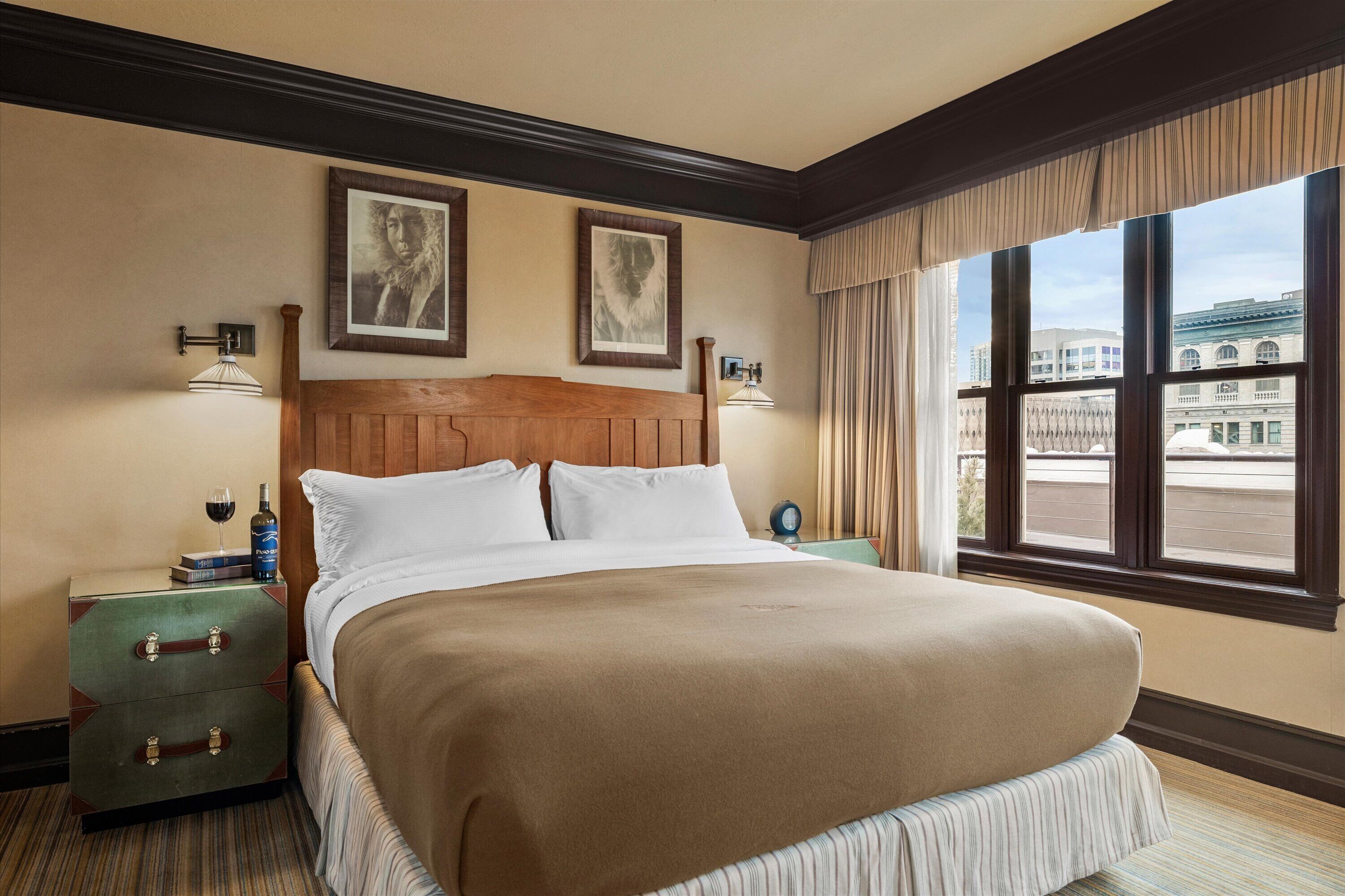A hotel room with a large bed, two framed pictures on the wall, a nightstand with a wine bottle, and a view of buildings through the window.