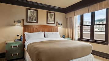 A hotel room with a large bed, two framed pictures on the wall, a nightstand with a wine bottle, and a view of buildings through the window.