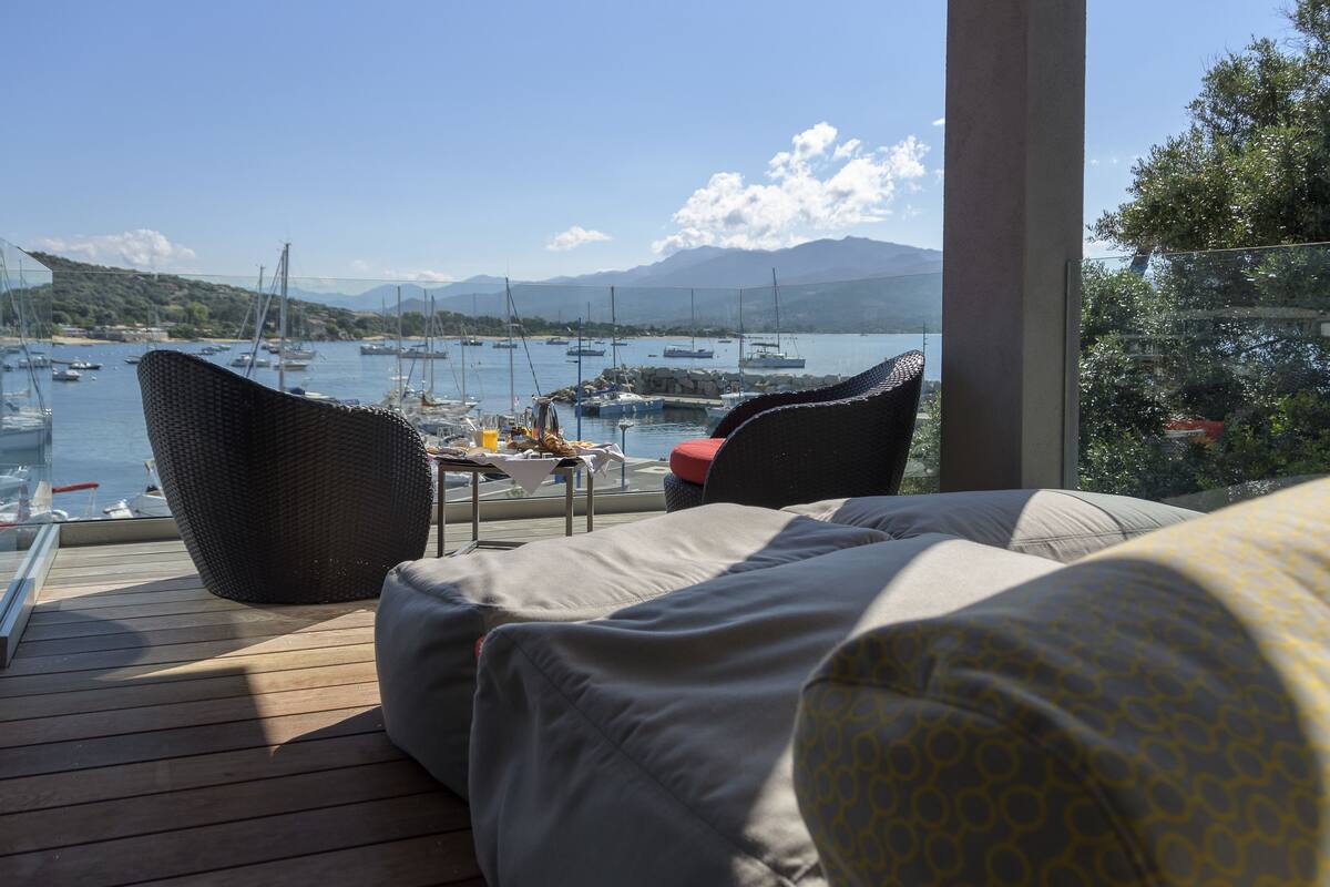 A balcony with wicker furniture, a table with a glass of orange juice, and a view of boats and mountains.