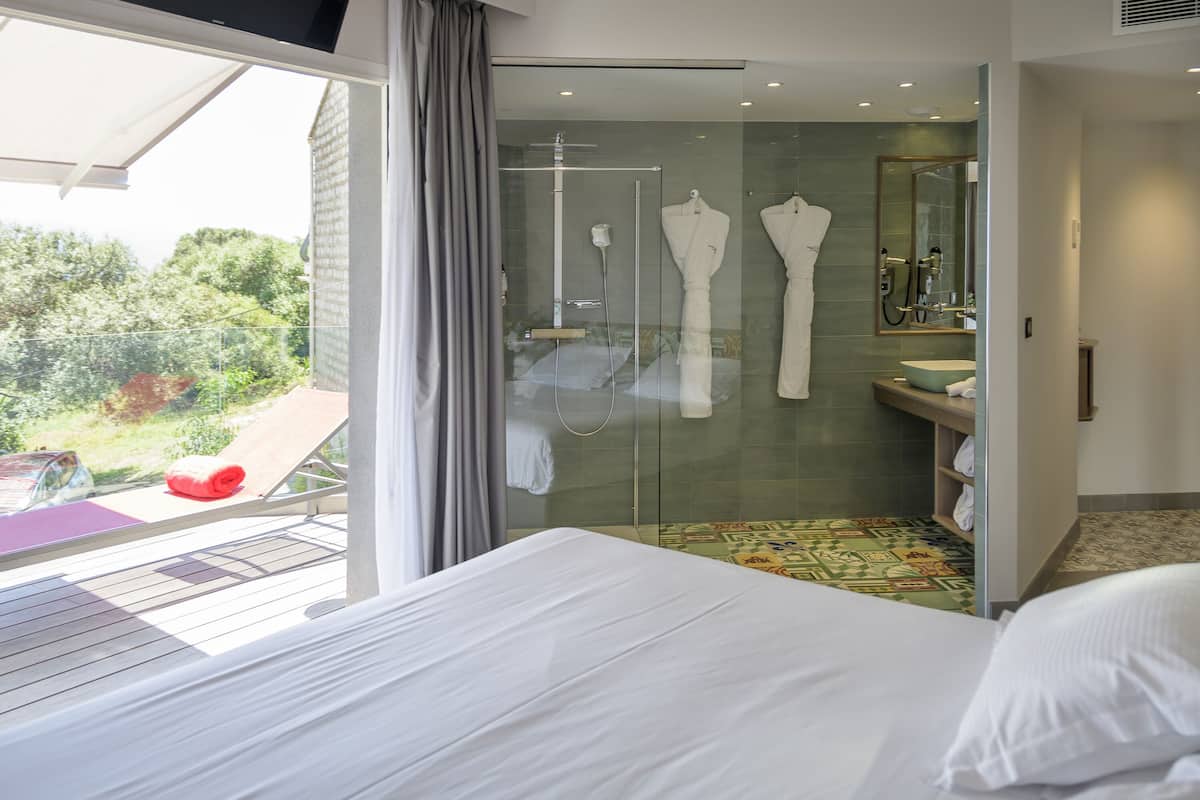 A hotel room with a large bed, a glass shower enclosure, a bathroom with a sink and mirror, and a balcony with a red chair.
