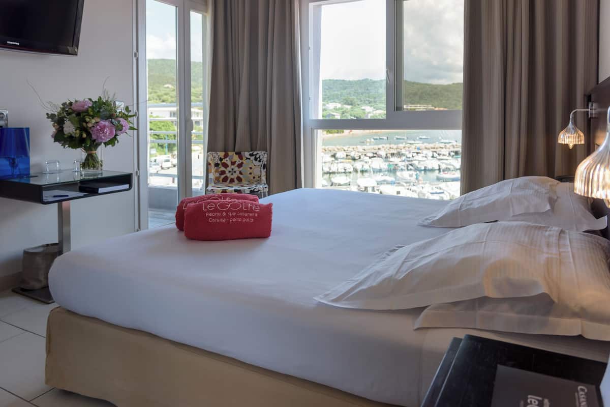 A hotel room with a bed, a desk with a TV, a chair, and a view of a marina.