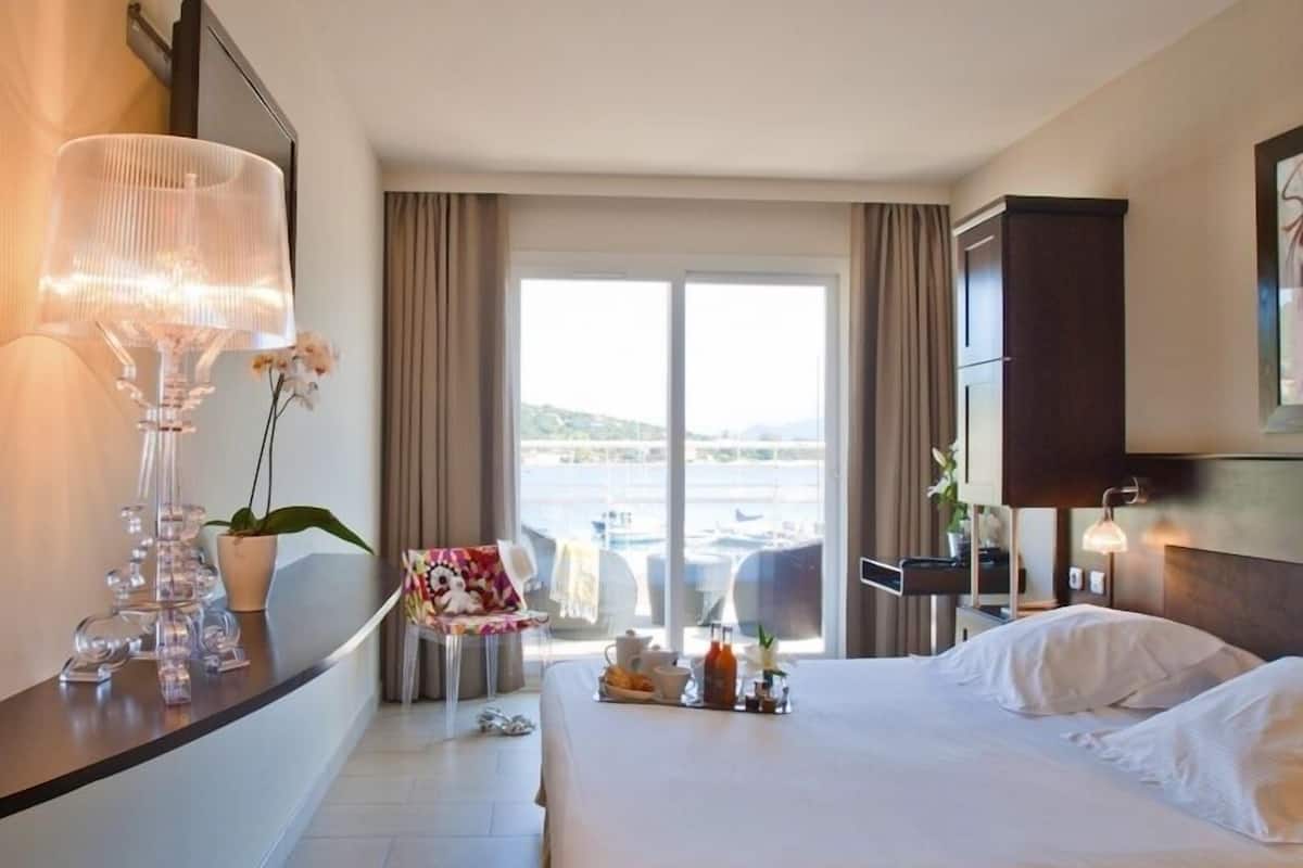 A hotel room with a large bed, a desk with a lamp, a chair, a small table with a plant, and a view of the sea through the window.