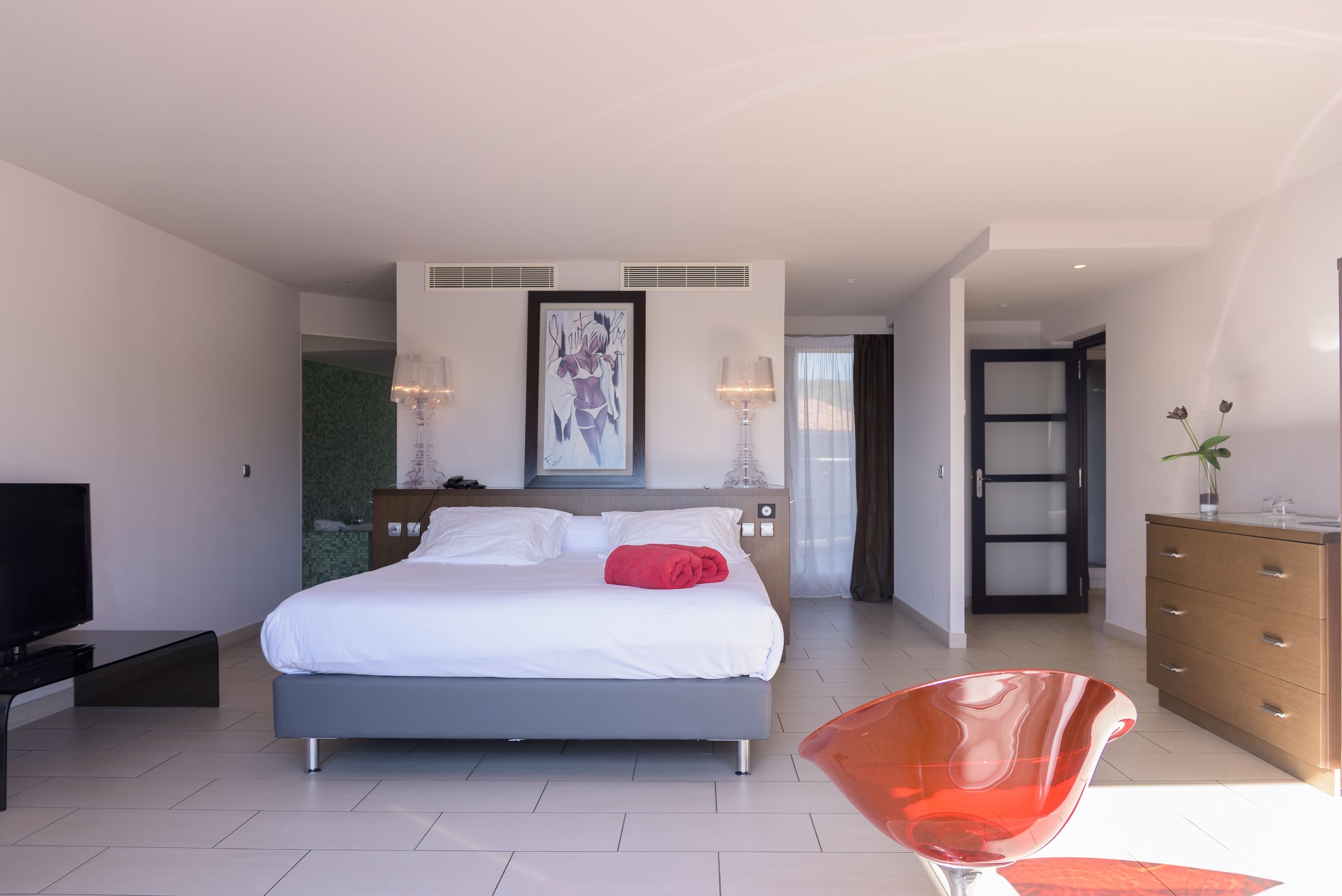 A modern hotel room with a large bed, a TV, a wooden dresser, and a bright red chair.