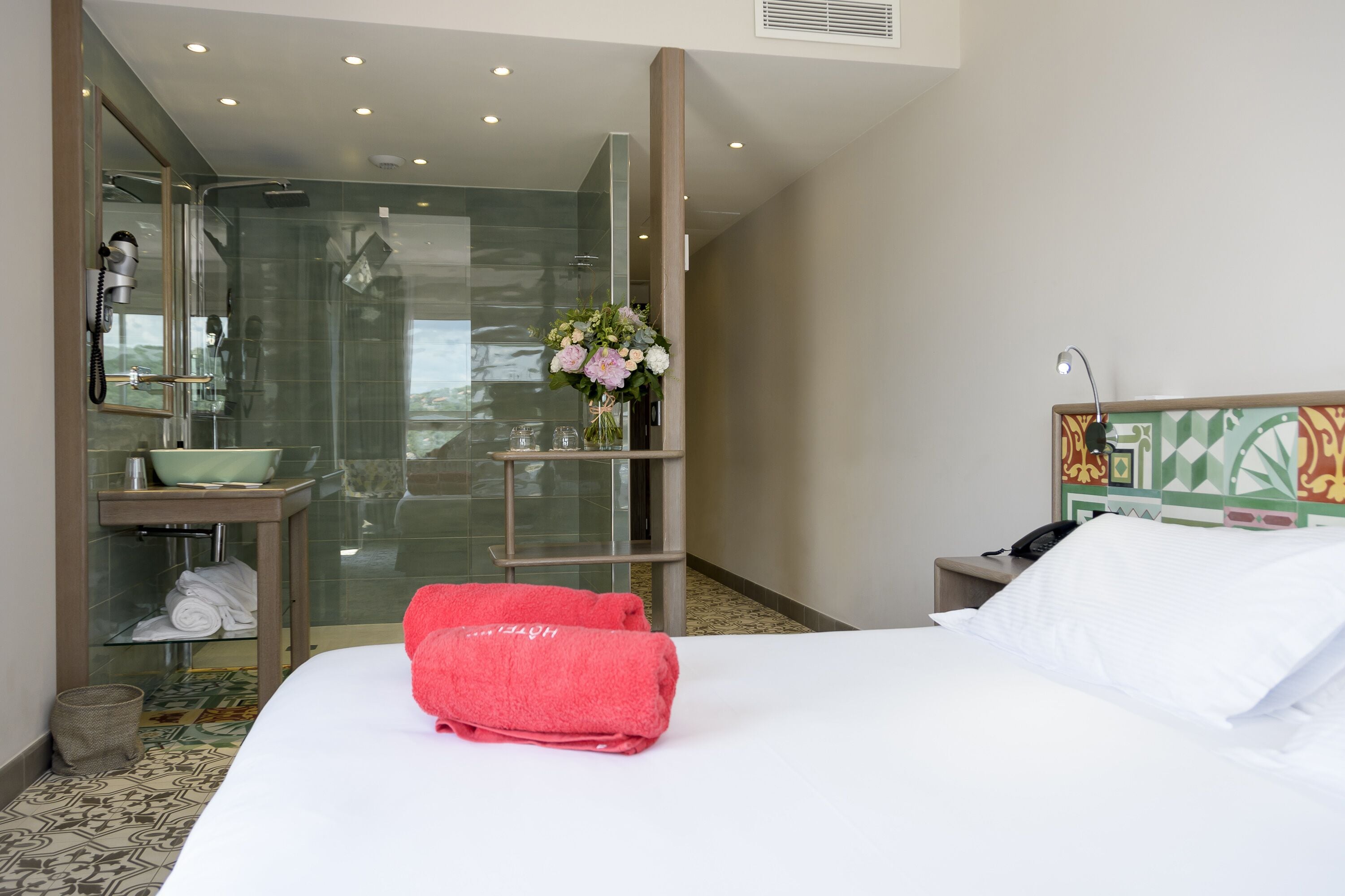 A hotel room with a bed, a glass-enclosed shower, a sink, and a vase with flowers.