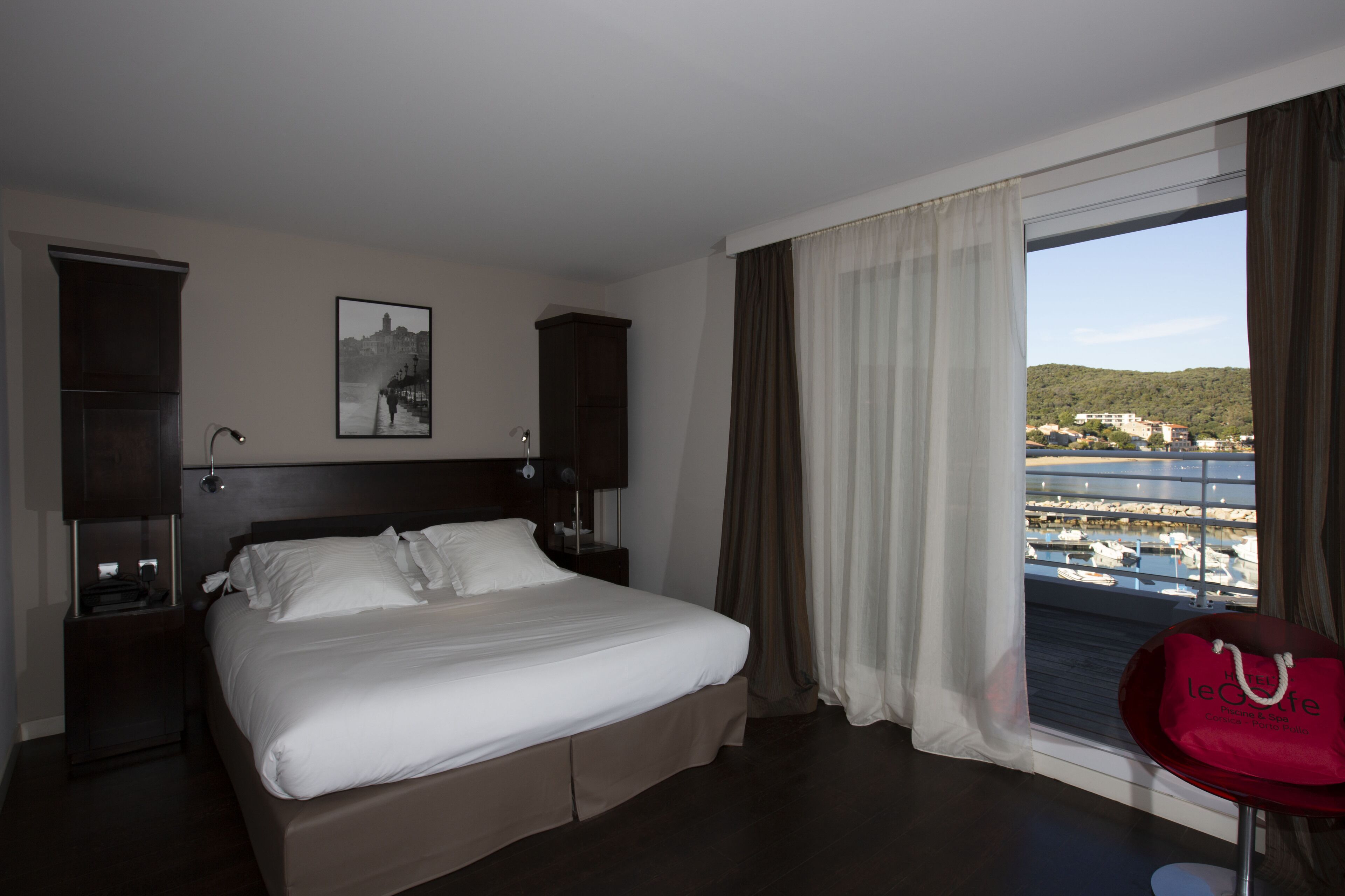 Habitació d'hotel amb llit, una tauleta de nit, una làmpada, una finestra amb cortines i vista al port esportiu.