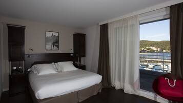 Habitació d'hotel amb llit, una tauleta de nit, una làmpada, una finestra amb cortines i vista al port esportiu.