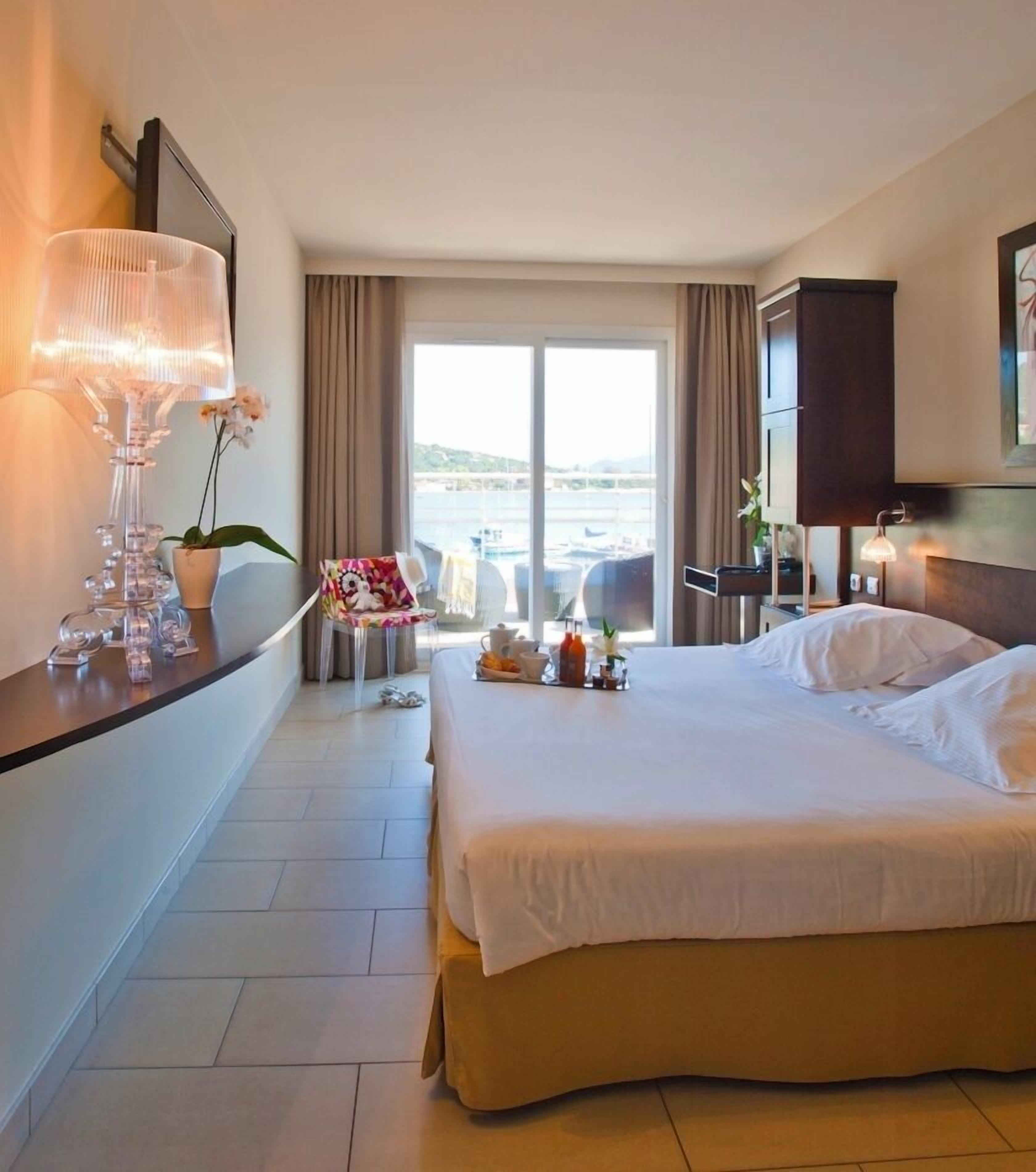 A hotel room with a large bed, a desk with a lamp, a chair, a small table with a plant, and a view of the sea through the window.