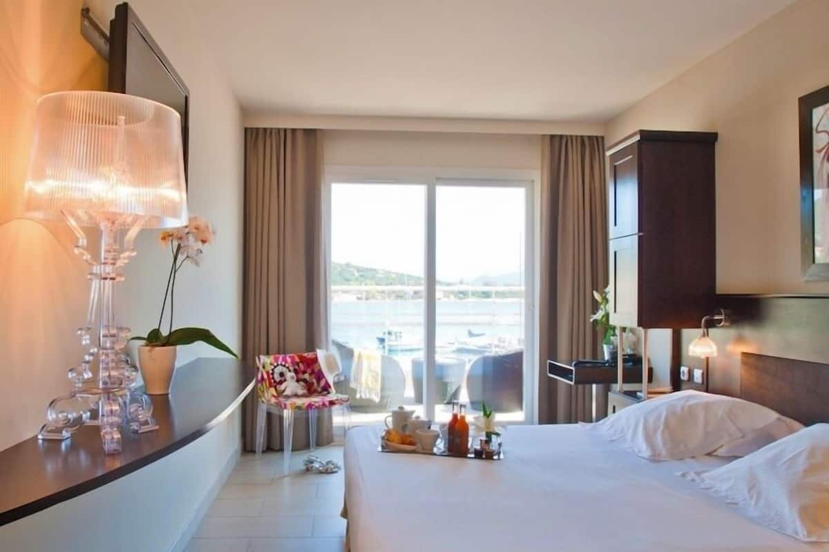 A hotel room with a large bed, a desk with a lamp, a chair, a small table with a plant, and a view of the sea through the window.
