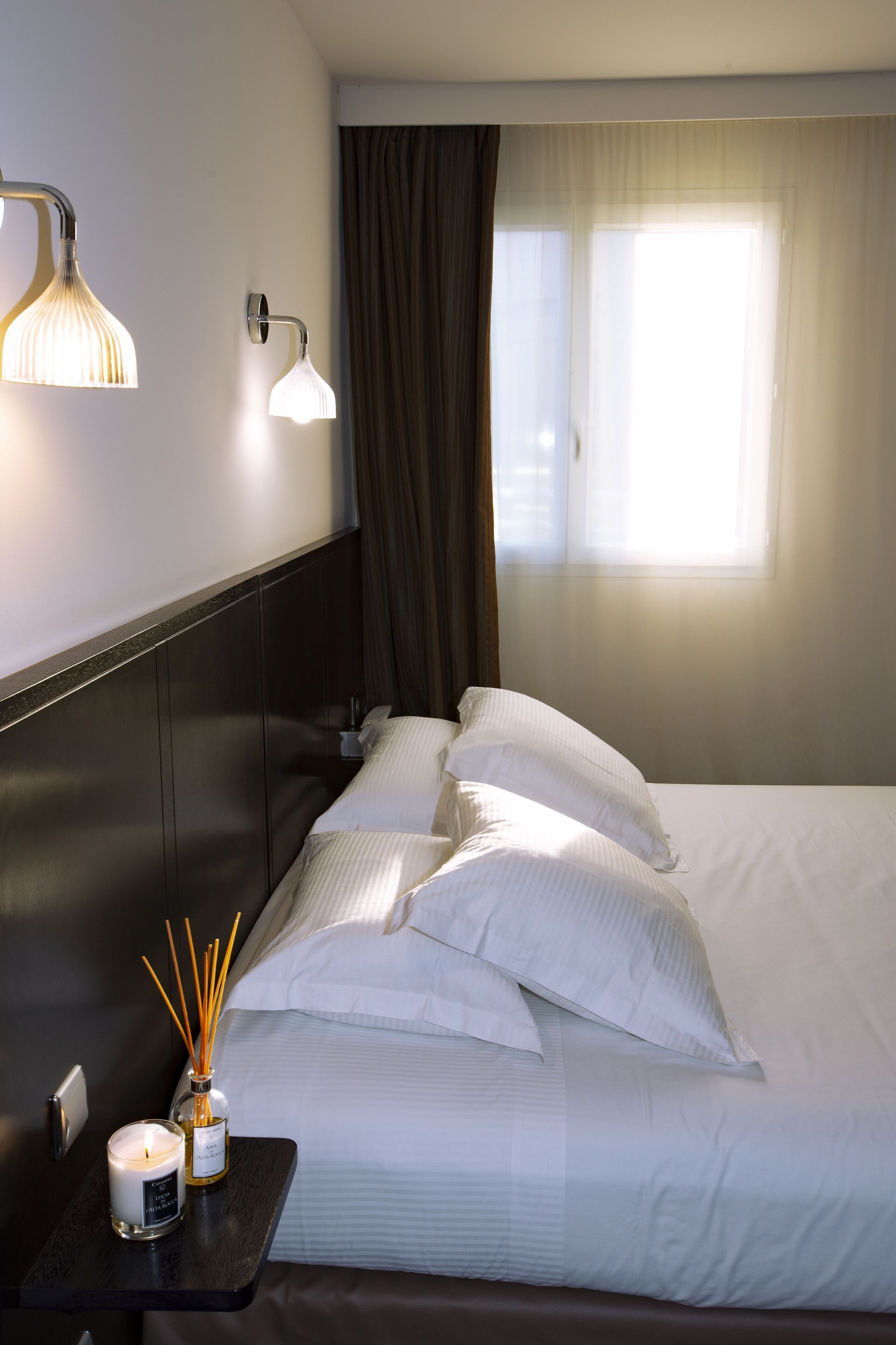A neatly made bed with white linens and pillows, a bedside table with a candle and reed diffuser, and a window with curtains.