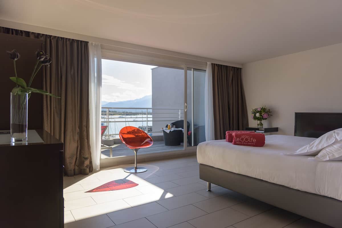 A hotel room with a bed, a red chair, a desk with a vase, and a view of the outdoors.