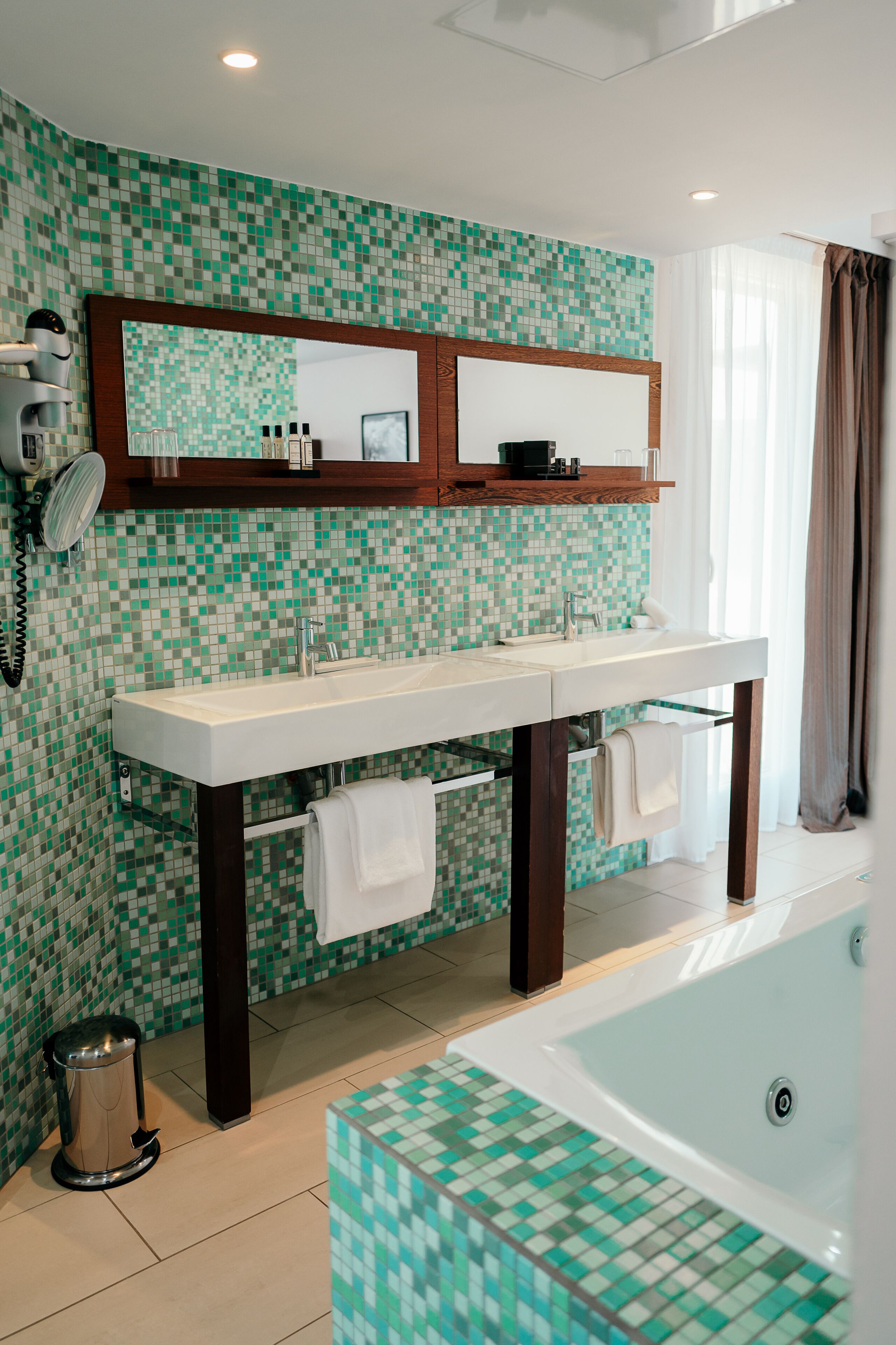 A bathroom with a tiled mosaic backsplash, a large mirror, a double sink, a hairdryer, a trash can, and a bathtub.