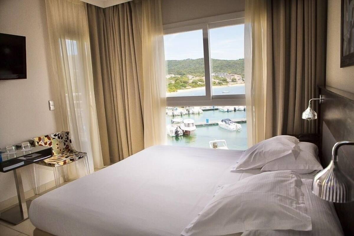 A hotel room with a bed, a desk with a chair, a TV, and a view of boats and a marina.