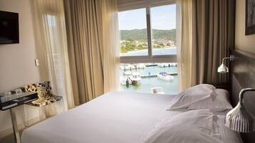 A hotel room with a bed, a desk with a chair, a TV, and a view of boats and a marina.