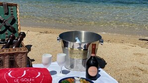 En picnicopsætning på en strand med et bord dækket til et måltid, inklusive skåle med mad, en flaske champagne og en halmhat.