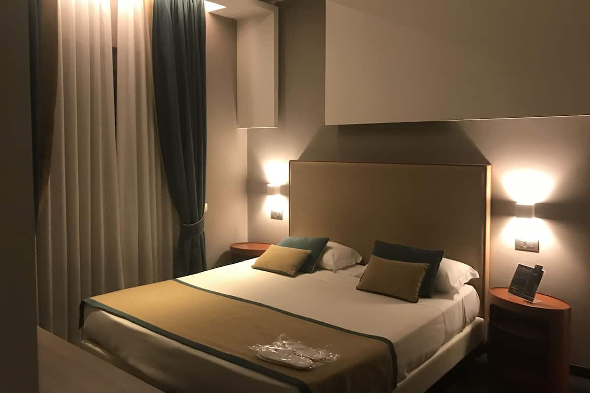 A hotel room with a large bed, two bedside tables, a nightstand, and a wall-mounted light fixture.