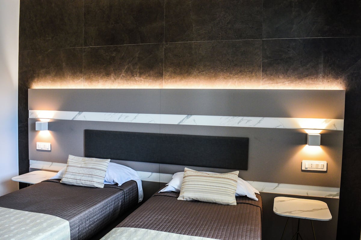 A hotel room with two beds, each with a headboard, bedside tables, and wall-mounted lighting.