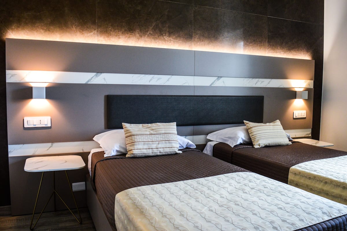 A hotel room with two beds, a small table, and wall-mounted lights.