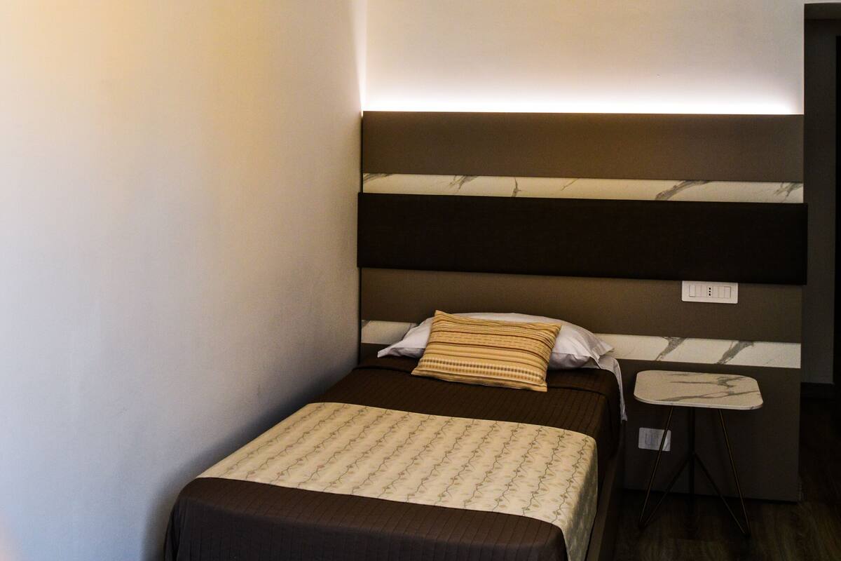 A small, modern hotel room with a single bed, a bedside table, and a nightstand.