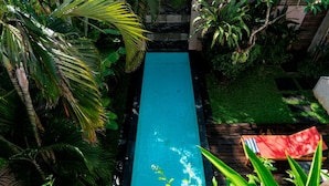 A rectangular pool with a wooden deck on one side, surrounded by lush greenery and palm trees. - Bali Island Villas and Spa Seminyak (Seminyak)