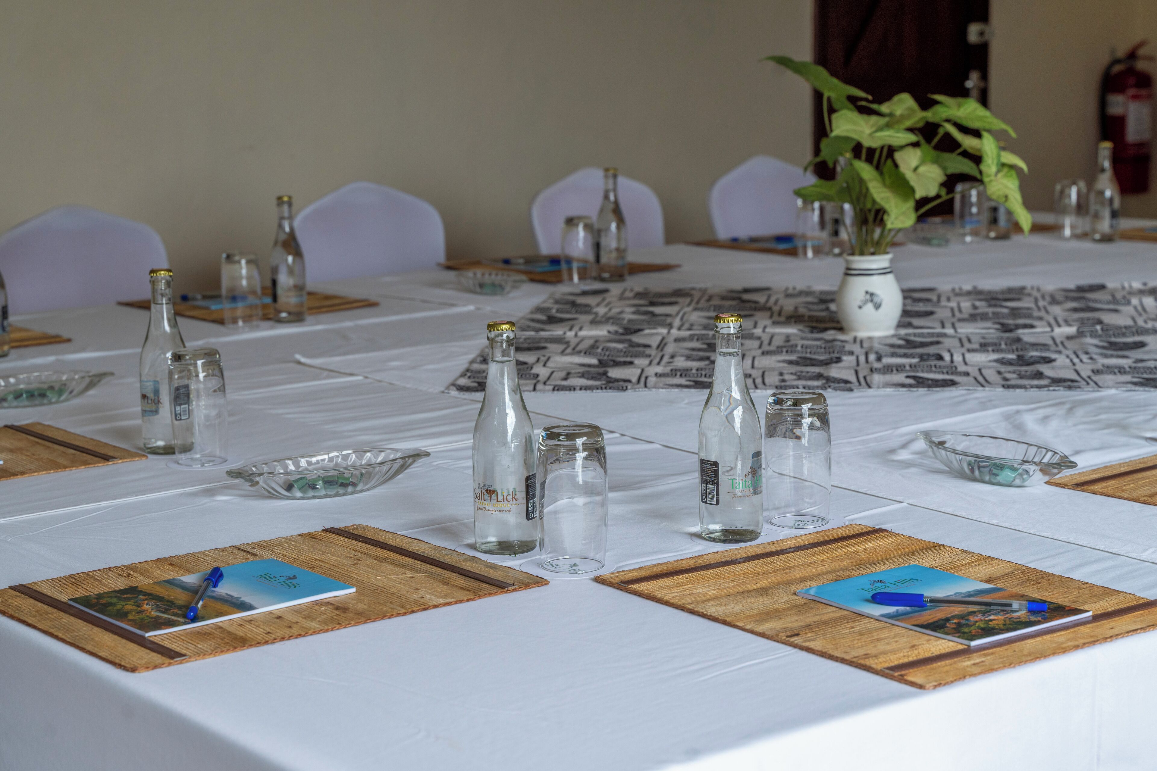 A table set for a meeting with glass bottles, a vase with a plant, and placemats with a book and pen.