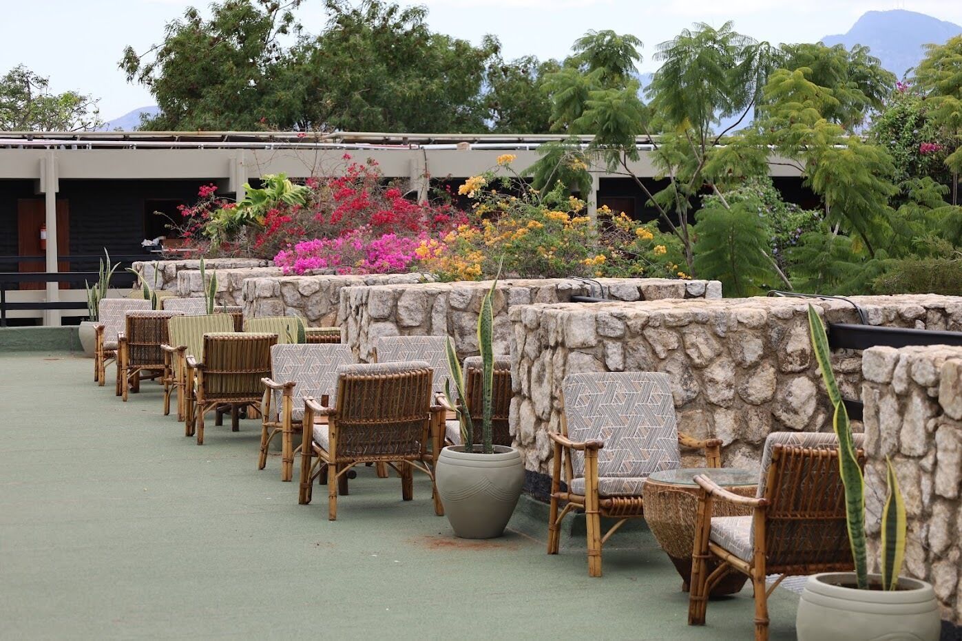 Outdoor seating with wicker chairs and planters, a stone wall, and a building in the background.
