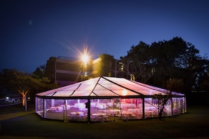 A large, illuminated tent with a glass roof, surrounded by trees and a building in the background.