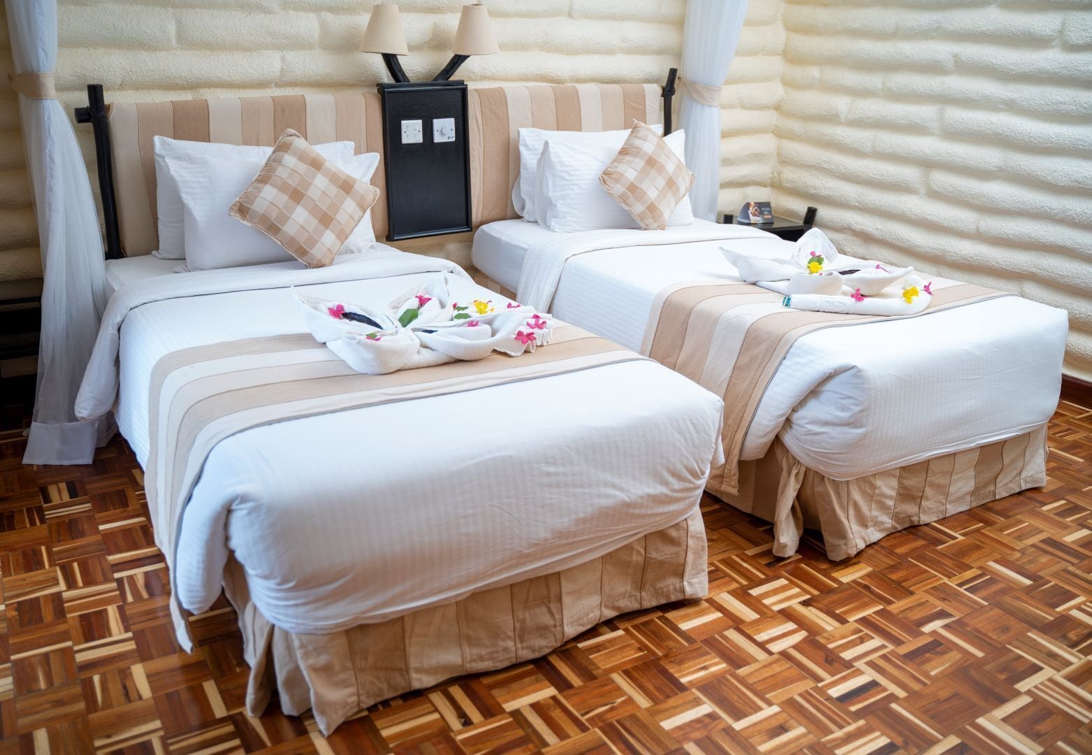 Quarto de hotel com duas camas, roupa de cama bege, piso de parquet de madeira e mesa de cabeceira com abajur.