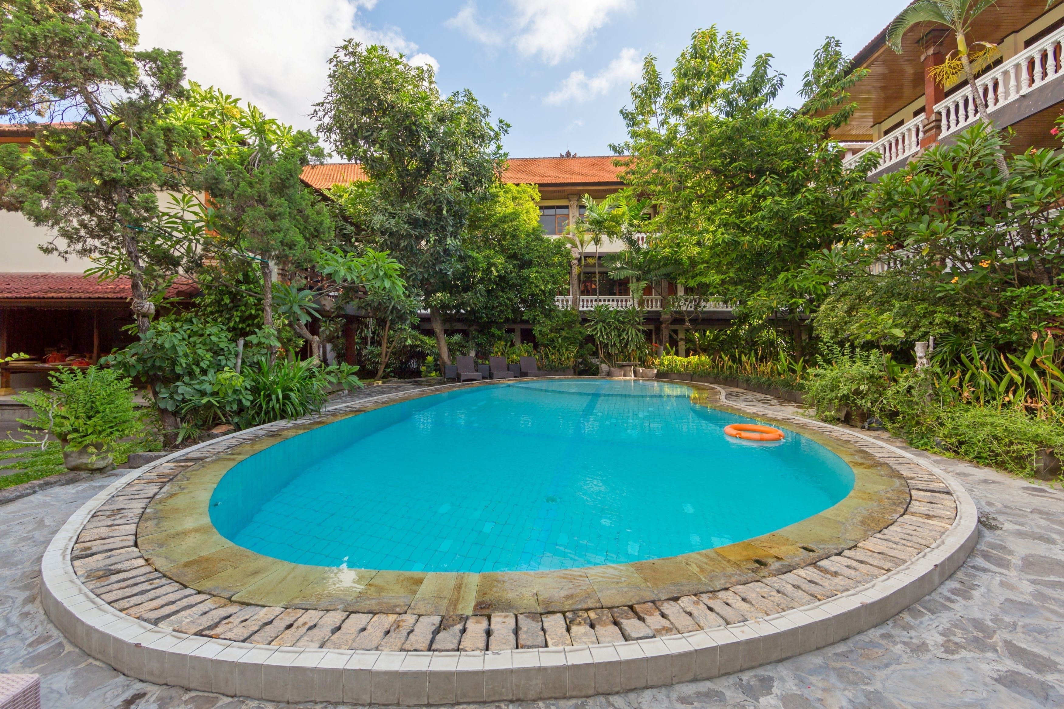 A circular pool with a tiled edge, surrounded by lush greenery and a building with a red roof in the background.