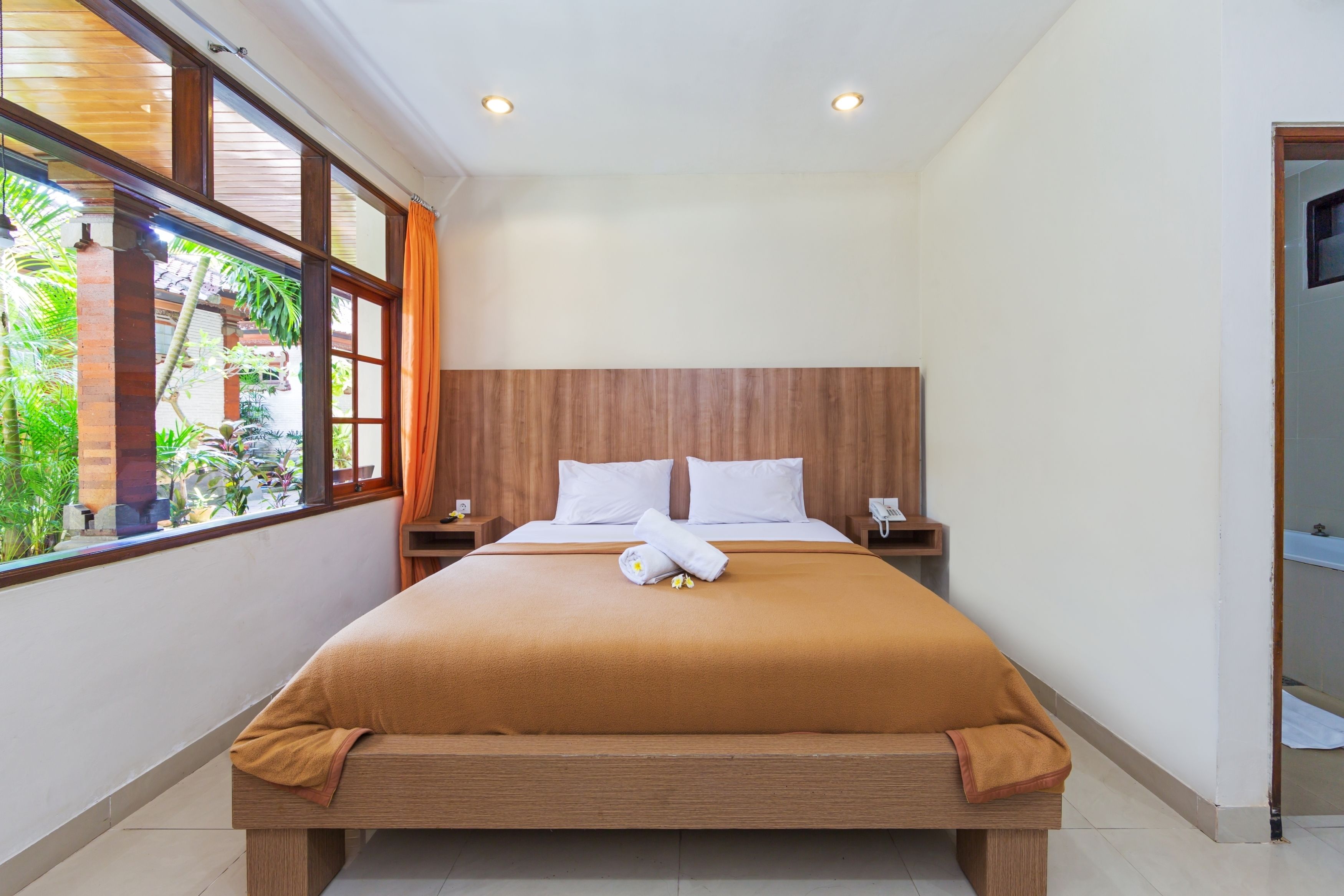 A neatly made bed with a brown bedspread, white pillows, and a rolled towel. There is a nightstand with a phone and a lamp. The room has large windows with curtains, and a view of greenery outside.