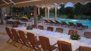 Uma sala de jantar com uma mesa comprida posta para uma refeição, rodeada de cadeiras, e uma piscina com espreguiçadeiras ao fundo.