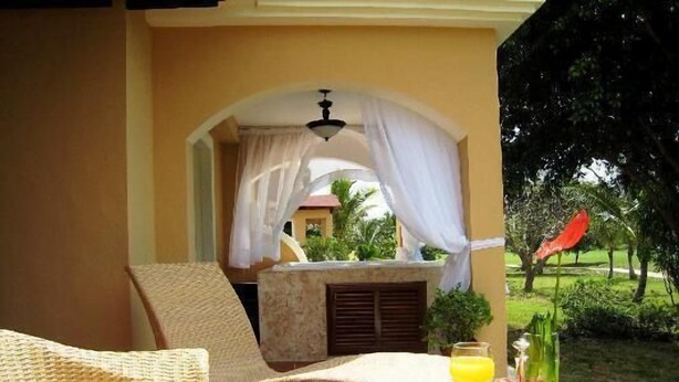A breakfast setup with a wicker table, a tray with food and drinks, and a view of a garden with a red umbrella. - The Golf Suites (Punta Cana)