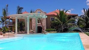 A swimming pool with a pergola, palm trees, and a building with a red roof. - The Golf Suites (Punta Cana)