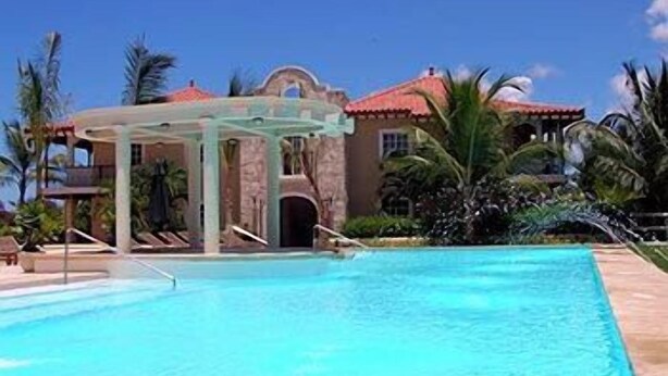 Uma piscina com pergolado, palmeiras e um edifício com telhado vermelho.
