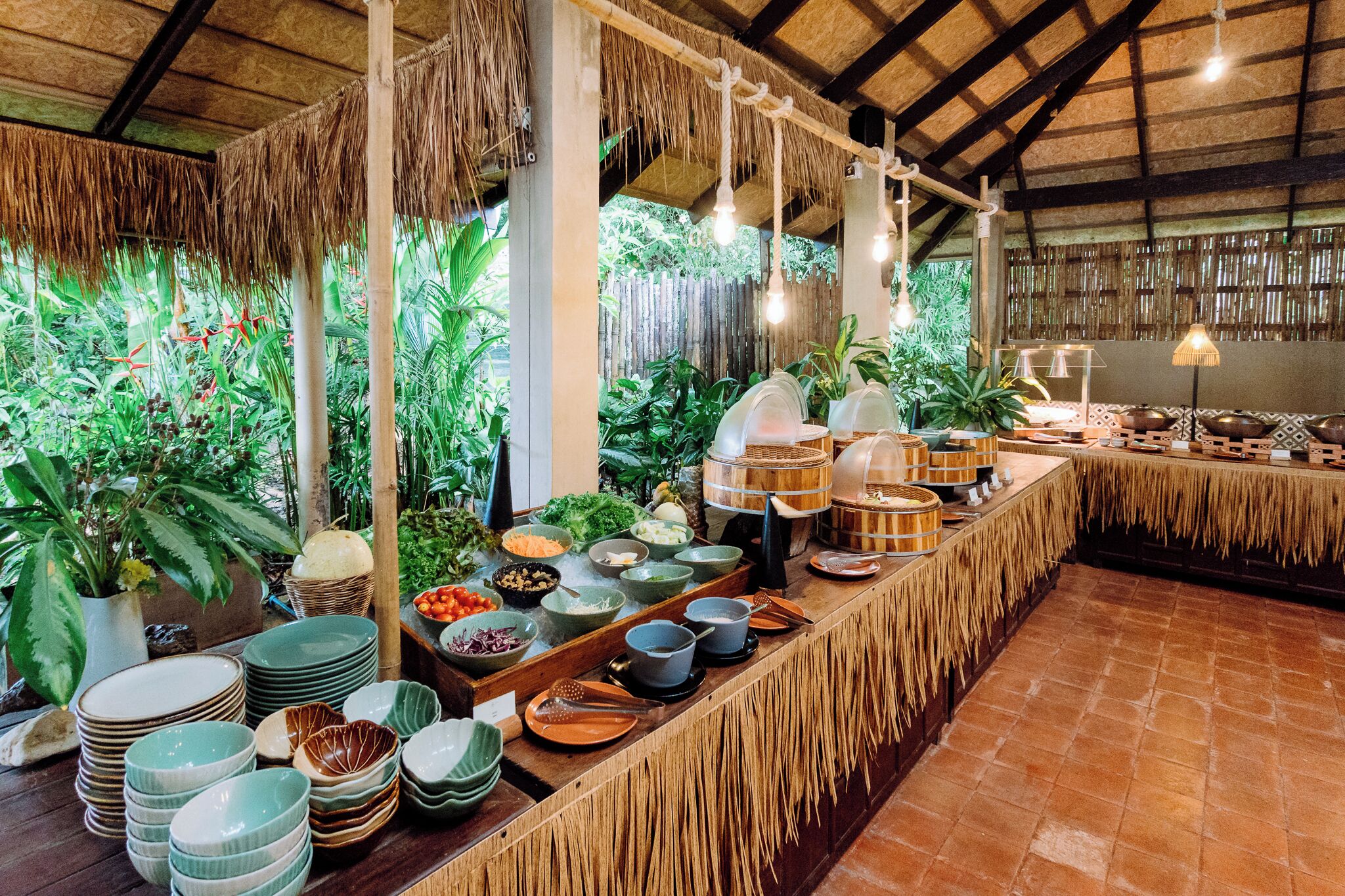 Un buffet proposant une variété de plats et d’ustensiles, entouré de plantes tropicales et surmonté d’un toit de chaume.