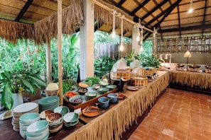 A buffet setup with various dishes and utensils, surrounded by tropical plants and a thatched roof.